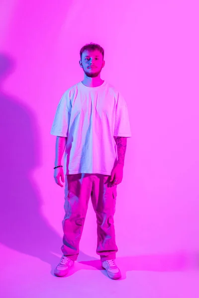 Stylish fashionable guy in a white T-shirt and pants with sneakers stands on a bright pink background with neon lights