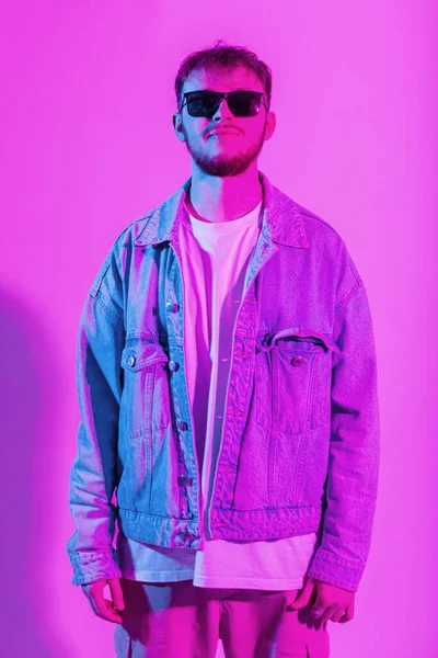 Trendy creative young handsome man with sunglasses in denim fashion jacket in the colorful studio with pink and blue light