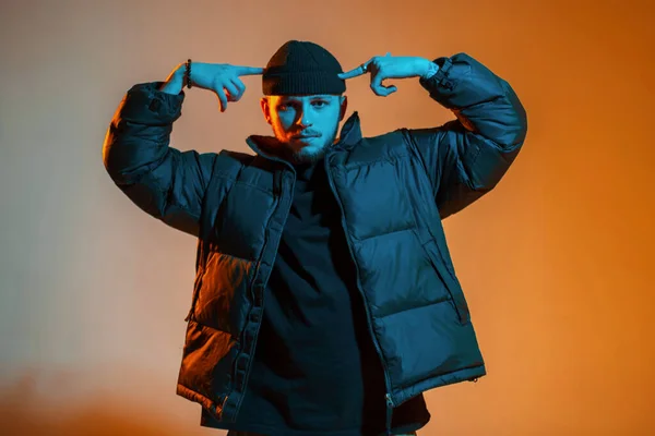 Cool hipster trendy man dancer in black outfit with cap and winter fashion black down jacket posing in creative studio with orange and neon light