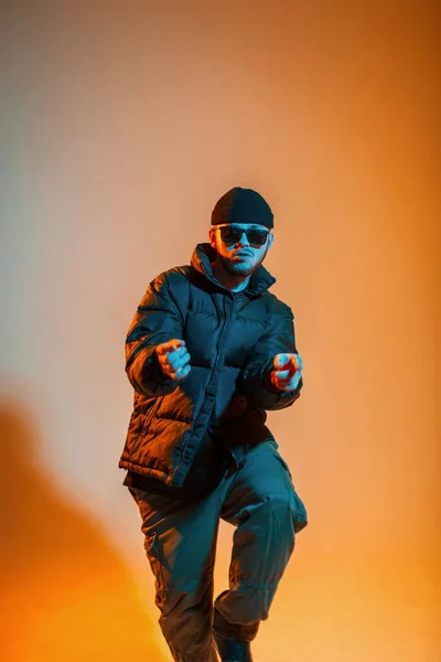 stock image Cool trendy stylish man dancer with fashion sunglasses in trendy black clothes with a hat and a down jacket is dancing in a creative color studio with warm yellow and cold blue light