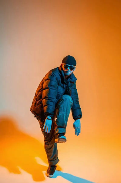 Stylish professional dancer guy in fashion black clothes with a hat, sunglasses, a down jacket, pants and sneakers is dancing in a creative color studio with orange and neon lights