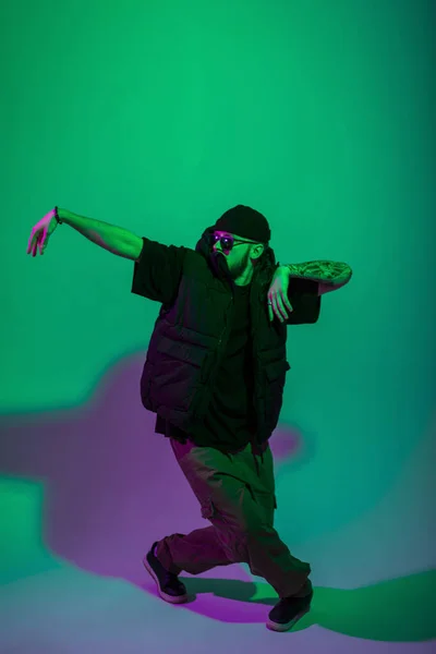 サングラスとスタイリッシュなベストと流行の黒の服の帽子を持つクールなファッションダンサーの男は緑と紫の光と創造的なスタジオで踊っています — ストック写真
