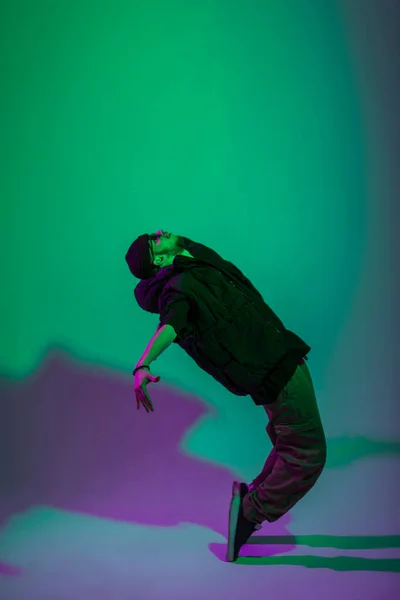 Fashionable handsome professional hip hop dancer man with a hat in a vest, jeans and sneakers dances in motion in a creative studio with green and purple light