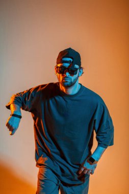 Güneş gözlüklü ve havalı tişörtlü havalı ciddi dansçı yaratıcı bir renk stüdyosunda turuncu ve neon ışıklı dans ediyor.