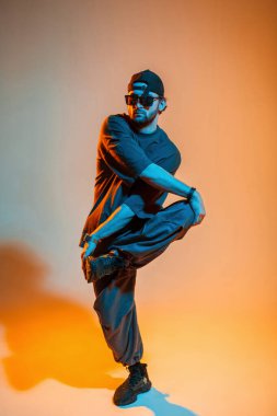 Havalı modaya uygun moda erkek model dansçı şapkalı, güneş gözlüklü, tişörtlü ve spor ayakkabılı turuncu ve neon ışıklı bir stüdyoda dans ediyor.