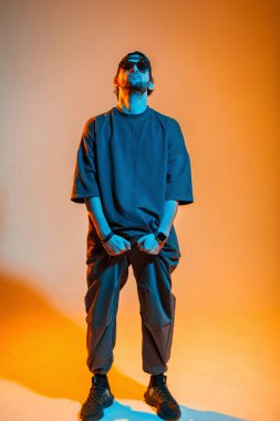 Moda kıyafetleri giymiş, güneş gözlüklü havalı profesyonel hip hop dansçısı pantolonunu tutuyor ve turuncu ve neon renkli bir stüdyoda poz veriyor.