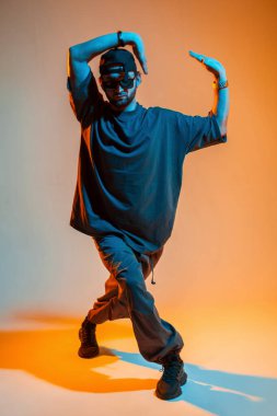 Güneş gözlüklü, şık ve şık bir erkek mankeni spor ayakkabılı şık bir şapkayla turuncu ve mavi ışıklı yaratıcı bir stüdyoda dans ediyor.