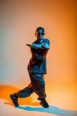 Şık, yakışıklı, güneş gözlüklü ve şapkalı profesyonel bir dansçı. Siyah elbiseli. Yaratıcı renk stüdyosunda turuncu ve neon ışıklı dans ediyor.