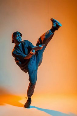 Havalı stil sahibi, yakışıklı, güneş gözlüklü ve tişörtlü şık şapkalı genç dansçı turuncu ve neon ışıklı bir stüdyoda dans ediyor.
