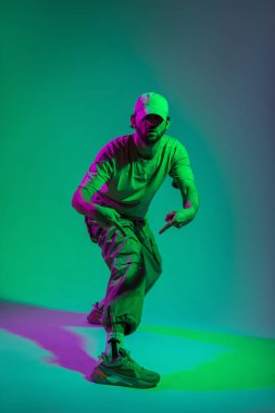 Havalı moda profesyonel dansçısı erkek manken şık kıyafetler içinde şık spor ayakkabılar giyiyor dans ediyor ve yaratıcı renkli stüdyoda yeşil ve pembe ışıklı poz veriyor.