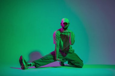 Moda kıyafetleri ve spor ayakkabılarıyla dans eden, yeşil ve pembe ışıklı bir stüdyoda oturan şık, yakışıklı, profesyonel bir dansçı.