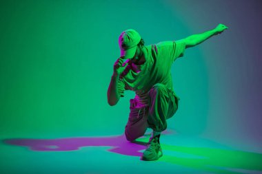 Moda kıyafetleri içinde şık, şık bir erkek model dansçı. Şapka, tişört, pantolon ve kot pantolon. Yaratıcı bir renk stüdyosunda yeşil ve mor ışık altında poz verip dans ediyorlar.