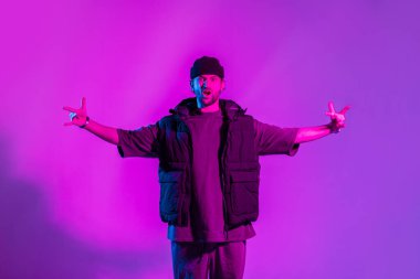 Şık giyinmiş, şapkalı ve yelekli komik dansçı stüdyoda yaratıcı pembe ve neon ışıkla duygusal poz veriyor.