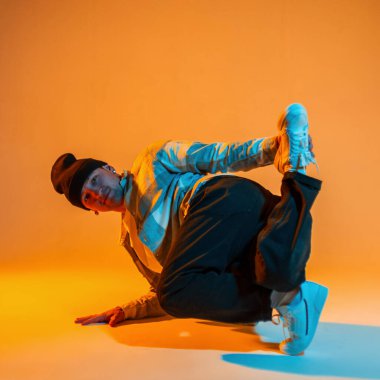 Profesyonel dansçı, şapkalı, ekose gömlekli ve spor ayakkabılı adam, yaratıcı bir renk stüdyosunda turuncu ve mavi ışıklı dans ediyor.