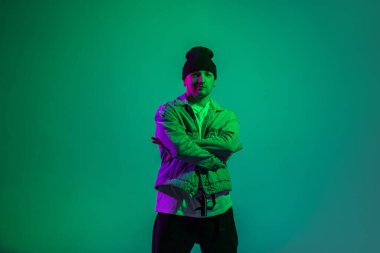 Havalı, şık break dansçı, modaya uygun kot elbiseli, şapkalı ve kot ceketli adam pembe ışıklı yeşil arka planda duruyor.