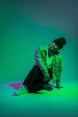 Yakışıklı, mutlu, modaya uygun kot elbiseli profesyonel dansçı stüdyoda yeşil ve mor ışıklı renkli bir arka planda oturuyor.