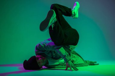 Havalı moda stilisti profesyonel break dansçı kot ceketli ve spor ayakkabılı mor ve yeşil ışıklı yaratıcı stüdyoda dans ediyor.