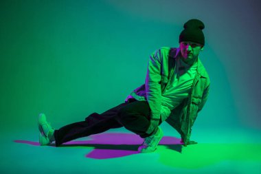 Moda sanatçısı B-boy adam şık kostüm giyip dans ediyor ve yaratıcı stüdyoda yeşil ve mor renkli pozlar veriyor.