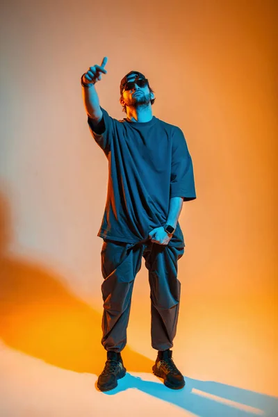 スタイリッシュなサングラスと流行の服のキャップを持つファッショナブルなハンサムな男が立っており オレンジと青の光と創造的なカラースタジオでポーズ — ストック写真