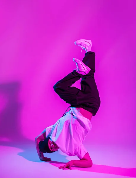 Stylish handsome b-boy dancer man in fashionable clothes with a hat, jeans, a t-shirt and sneakers stands on his arm and dances in a creative studio with pink and neon lights