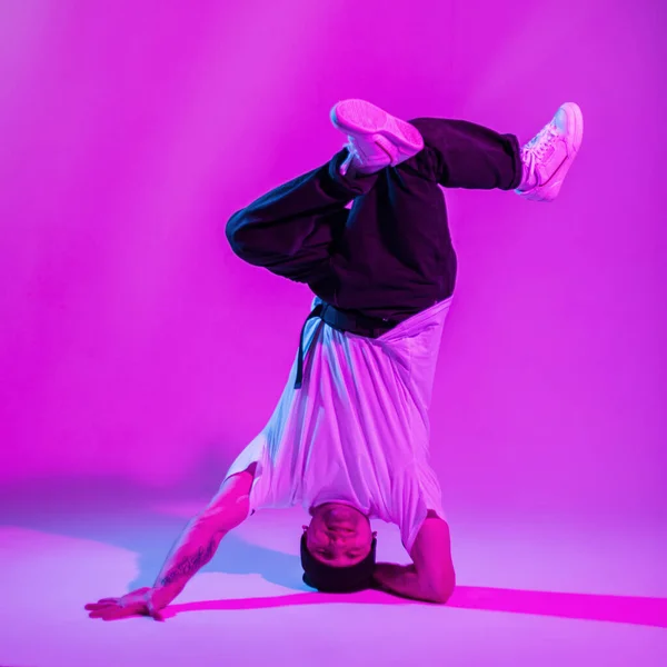 stock image Cool fashionable professional male breakdancer in trendy fashion clothes with hat, t-shirt and sneakers stands on head and dances in a creative color studio with pink and neon lights