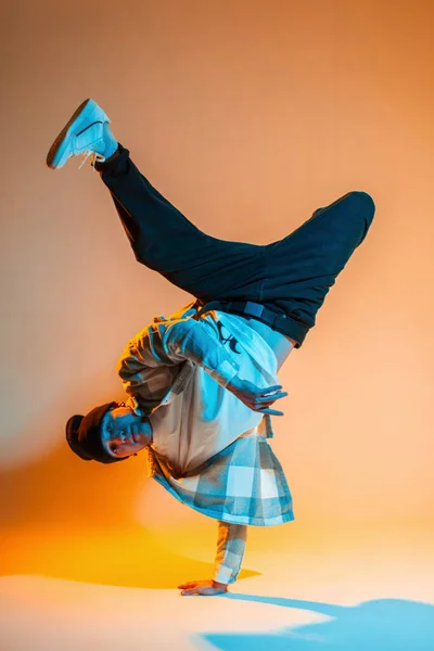 stock image Stylish handsome dancer breakdancer b-boy man in fashionable clothes with a shirt and sneakers dances and one hand freeze in a creative color studio with warm orange and cold blue light