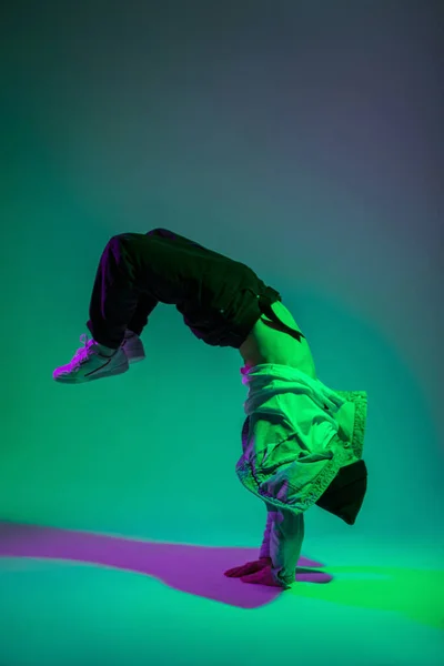 Cool stylish young professional dancer man b-boy in trendy fashion denim clothes with sneakers is dancing and freeze on hands in a creative color studio with pink and green light. Dance in motion
