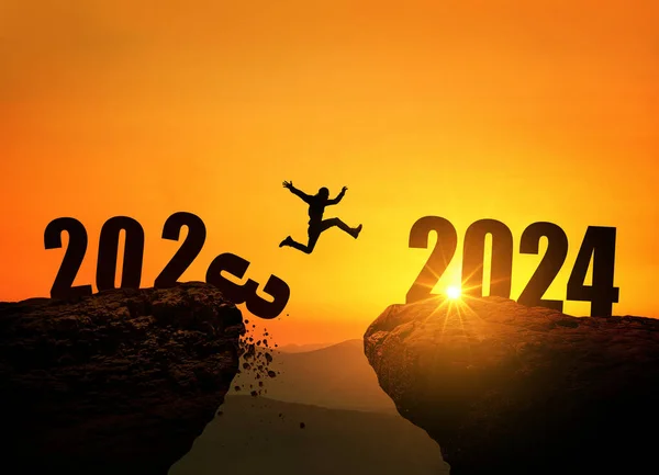 Man jumping on cliff 2024 over the precipice with stones at amazing sunset. New Year's concept. 2023 falls into the abyss. Welcome 2024. People enters the year 2024, creative idea.