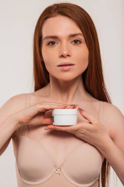 Bej sütyenli güzel, genç, kızıl saçlı bir kız beyaz arka planda beyaz bir kavanozda krema tutuyor. Güzellik ve cilt bakımı