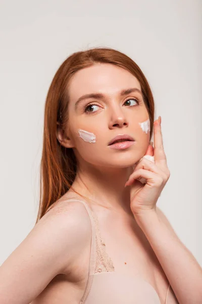 stock image Red-haired beautiful fresh young woman applies cream on the skin of the face in the studio on a white background. Beauty and skin care