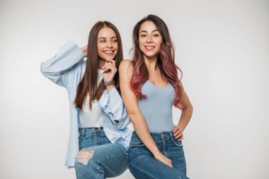Modadan güzel, genç, mutlu, şık kot elbiseli iki kız arkadaş stüdyoda beyaz bir arka planda poz veriyor ve eğleniyorlar.