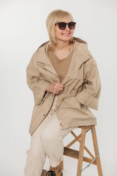 Happy fashion senior blonde woman with a smile with vintage sunglasses in fashionable beige clothes with a jacket sits on a wooden chair on a white background