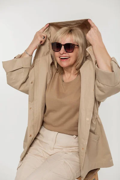 Funny happy fashion senior woman with cool vintage sunglasses in trendy fashion beige outfit with a jacket puts on a hood and has fun on a white background