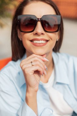 Neşeli bir kızın mutlu sokak portresi zarif, klasik güneş gözlüklü, güneşli bir bahar gününde oturur ve dinlenir.