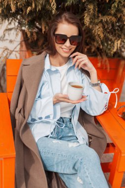 Güzel bir gülümsemesi olan şık güneş gözlüklü, modaya uygun kıyafetleri olan, bir kafede oturan ve güneşli bir bahar gününde çay içen oldukça mutlu bir kadın. Bahar geldi.