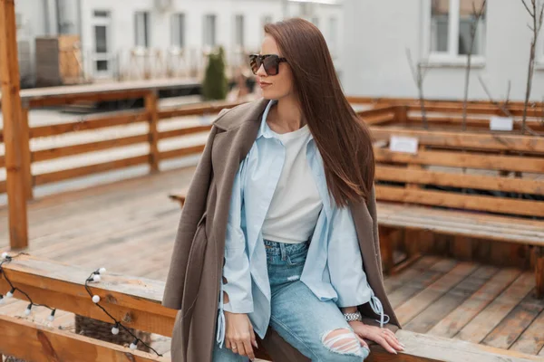 穿着时髦休闲装 穿着时髦外套 衬衫和牛仔裤的漂亮时髦姑娘 戴着老式太阳镜坐在街上的木制篱笆上休息 — 图库照片