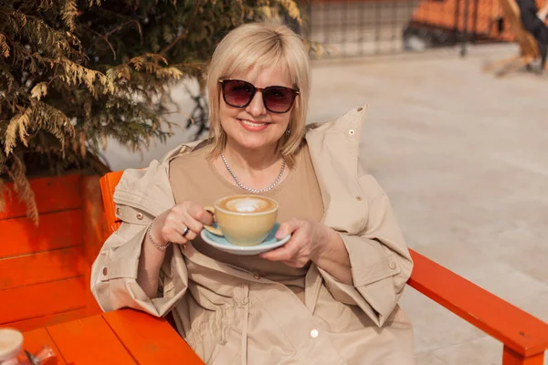 美丽而快乐的老妇人 戴着墨镜 穿着时髦的米黄色衣服 笑着坐在户外咖啡馆里 在一个阳光灿烂的春日里喝咖啡 — 图库照片
