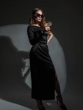 Havalı güneş gözlüklü, şık, topuklu siyah şık elbiseli güzel bir kadın bir partide bir bardak şampanya tutuyor. Hanımefendi bir olayı kutluyor.