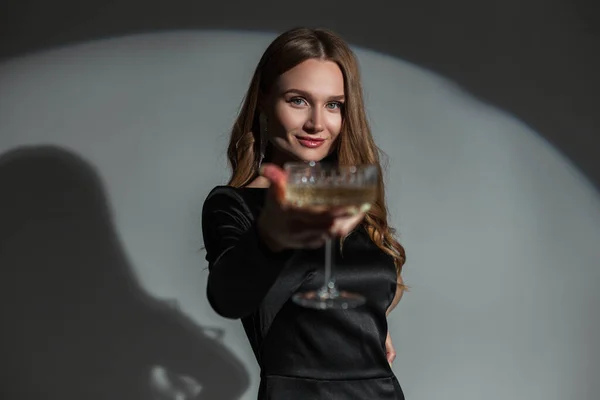 Stock image Beautiful happy stylish chic woman with a smile in a fashionable black elegant dress raises a glass of champagne on a dark background in the studio. Pretty lady at party