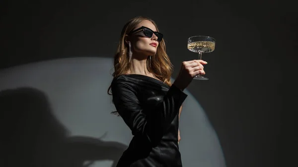 stock image Beautiful elegant young fashion woman with sunglasses in a trendy black dress with a vintage glass of champagne on a dark background in the studio. Lady celebrating a birthday at a party