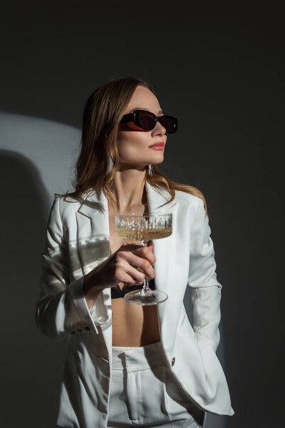 Cool fashion pretty chic woman with vintage sunglasses in a fashionable white suit with a glass of champagne on a dark background at a party