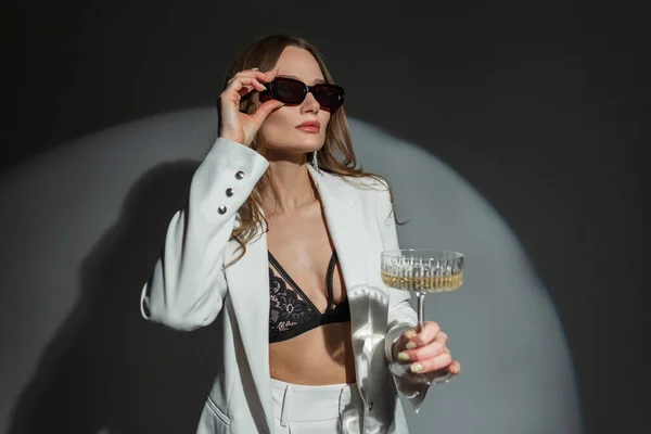 stock image Successful beautiful sexy woman in fashion white suit with blazer and lace bra holds champagne glass and puts on sunglasses on dark background in studio. Pretty chic lady celebration event