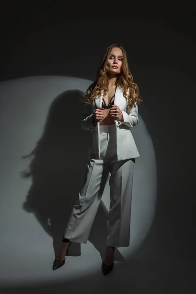 stock image Stylish beautiful elegant lady in a fashionable white suit with a bra and heels posing on a dark background with a circular light