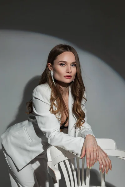 stock image Beautiful fashionable lady in a white stylish suit stands near a white chair on a dark background in the studio at a party