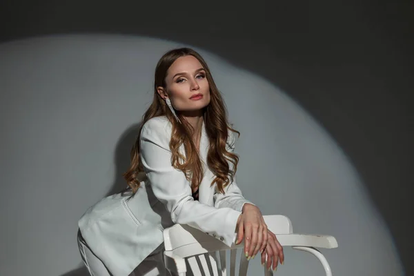 stock image Beautiful stylish successful business girl in an elegant white suit with blazer stands and poses near a white chair in the studio. Pretty chic lady at party