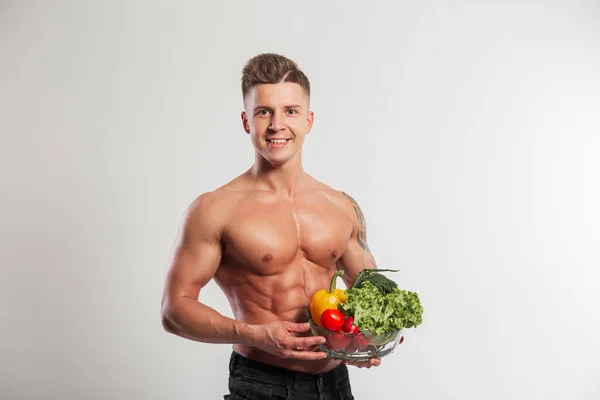 Happy handsome sports vegan athlete man with hairstyle with muscular handsome body holding glass bowl with vegetables on white background in studio