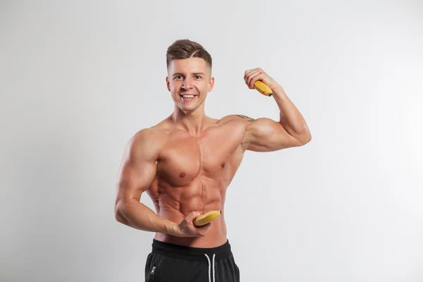 Funky handsome happy strong athletic man with muscular body holding bananas and showing biceps on white background in studio. Healthy raw nutrition and natural vitamins Potassium