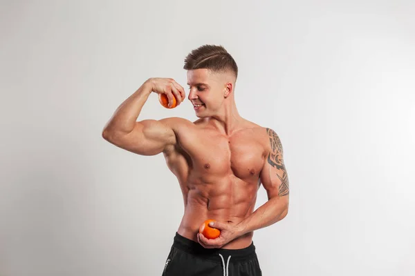 オレンジと筋肉質の体を持つ幸せなハンサムなフィットネストレーナーの男は スタジオで白い背景に両足を示しています 健康食品 運動やビタミン — ストック写真