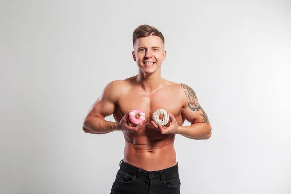Happy funny fitness muscular man with naked torso holding donuts on white background. Sports, diet and sweets