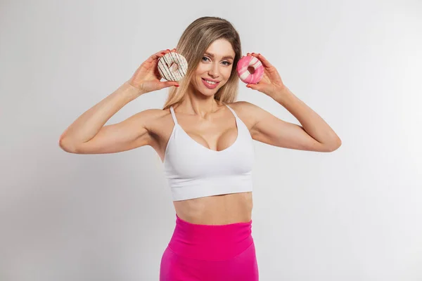 Beautiful happy young fitness lady with slim body in white top and pink leggings holding donuts on white background
